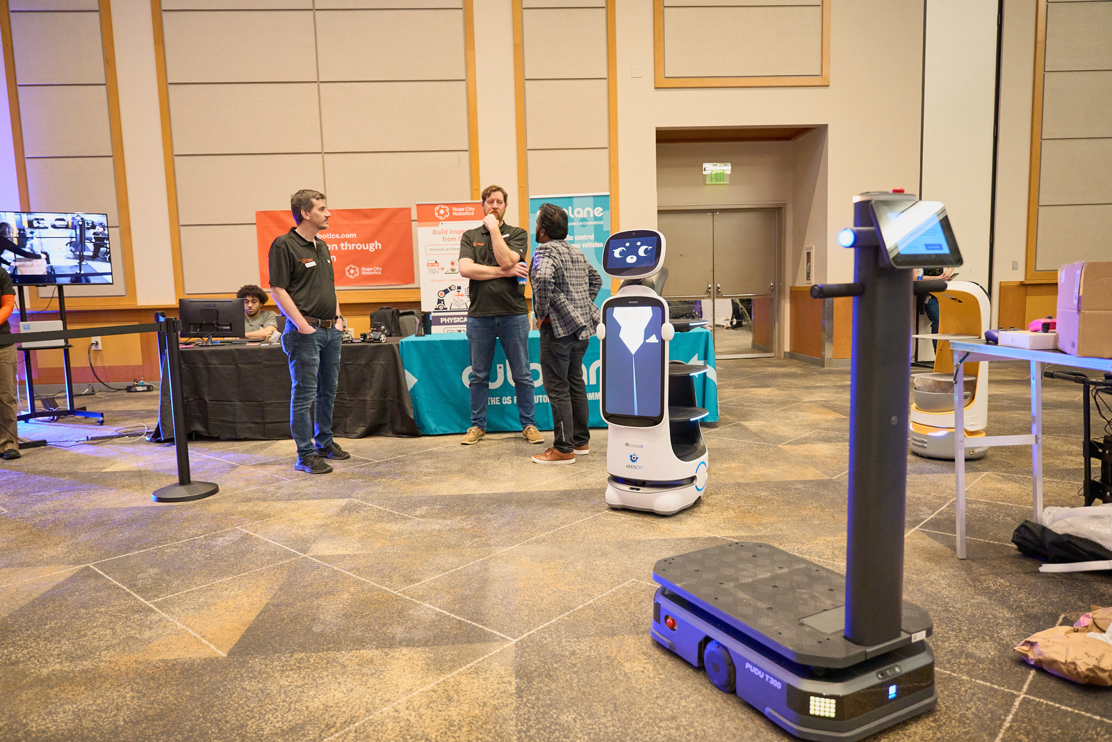 Wide view of the Silicon Forest Tech Summit 2026 event hall, showing the Rose City Robotics booth banner, the Autolane booth, a Bellabot service robot, and a Pudu cleaning robot, with two RCR engineers in conversation in the background.